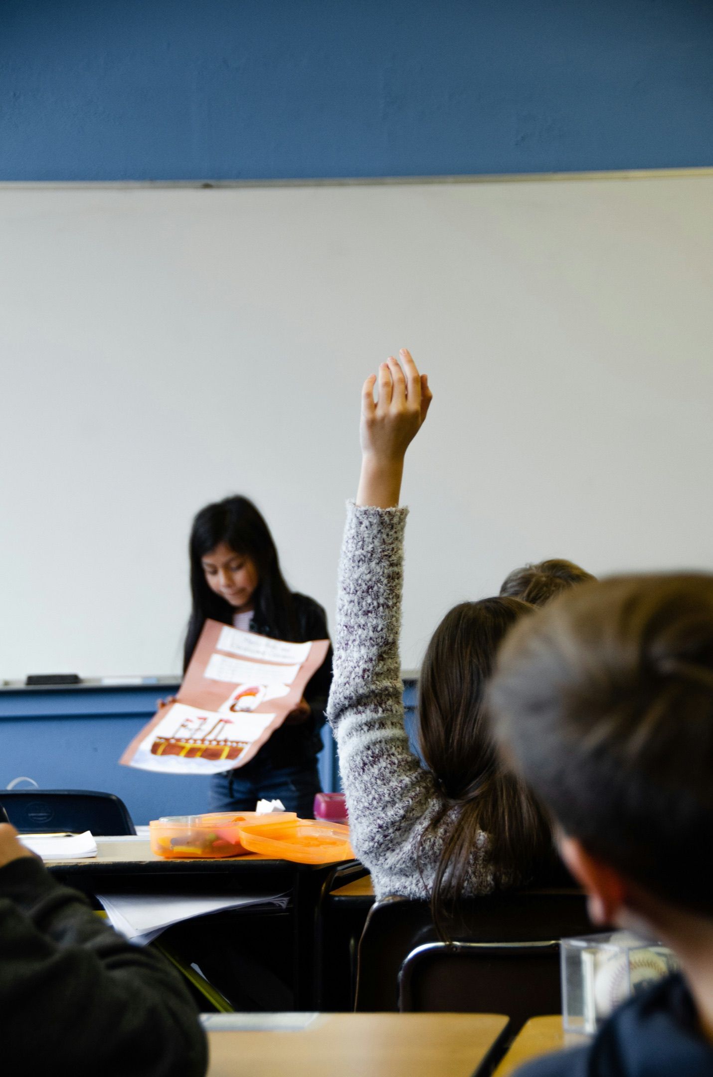 A student raises her hand while another student presents at the front of the classroom.