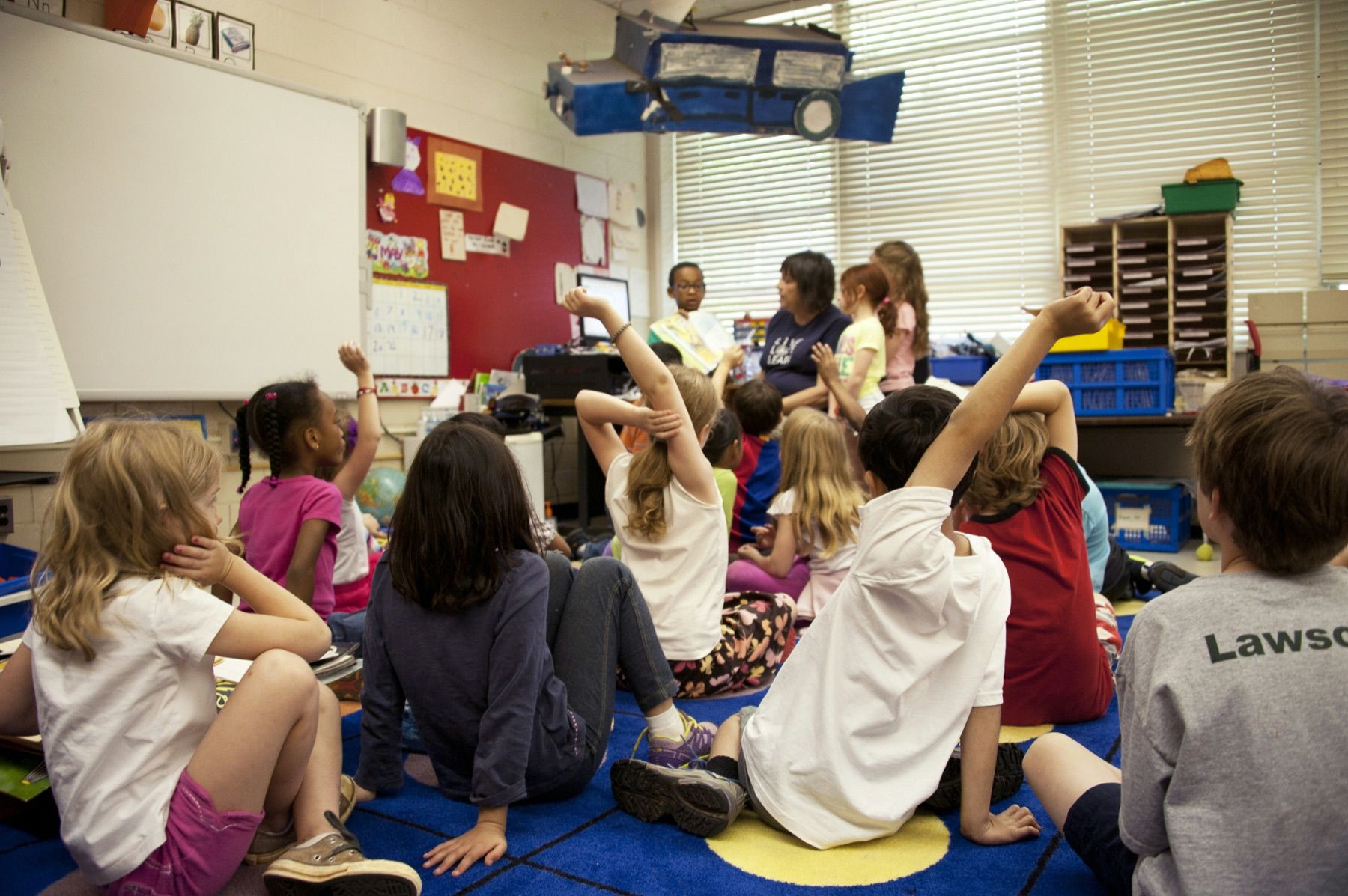Elementary students sit together on a classroom rug and raise their hands during a group reading activity.