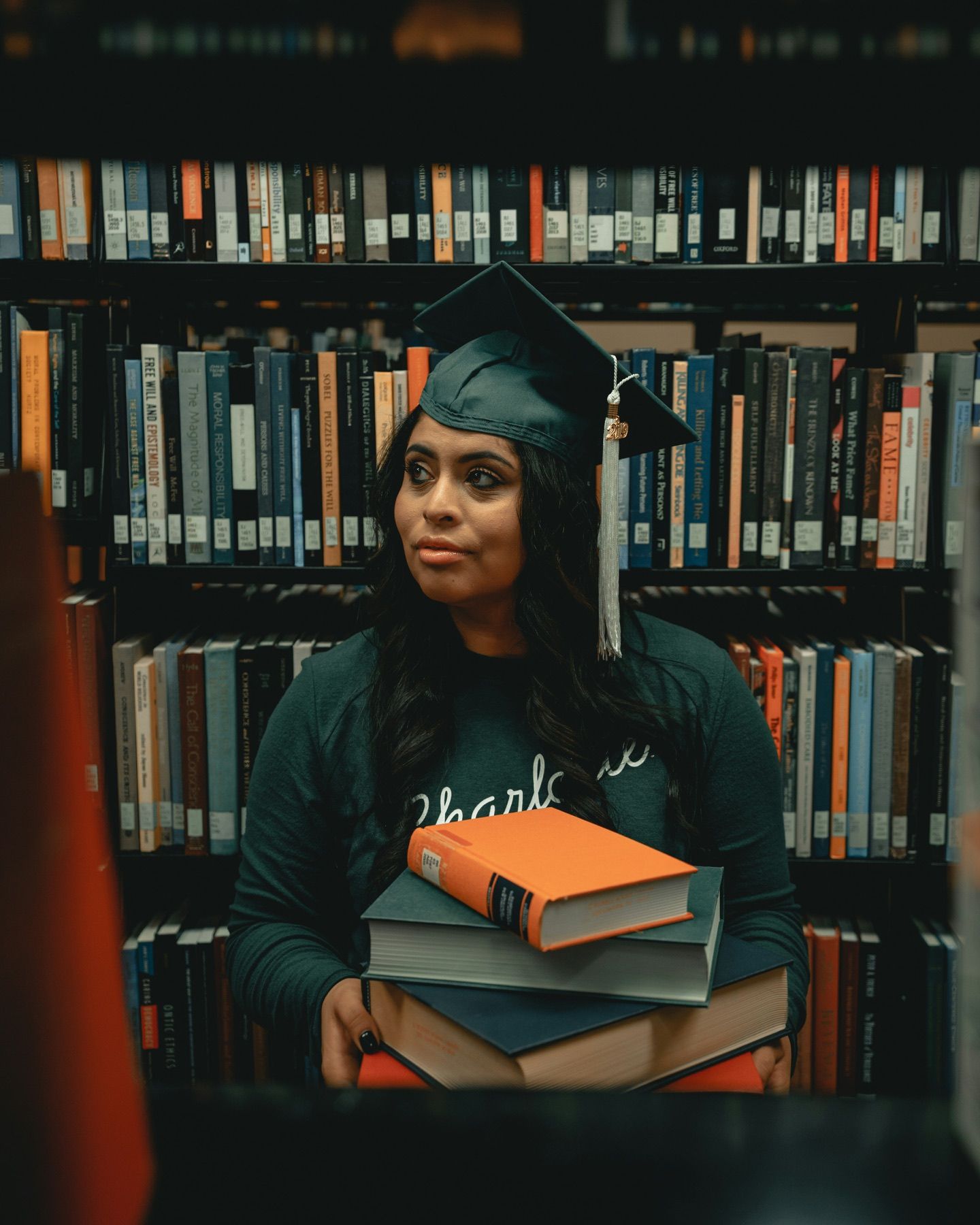 A graduate in a cap holds a stack of books between library shelves.