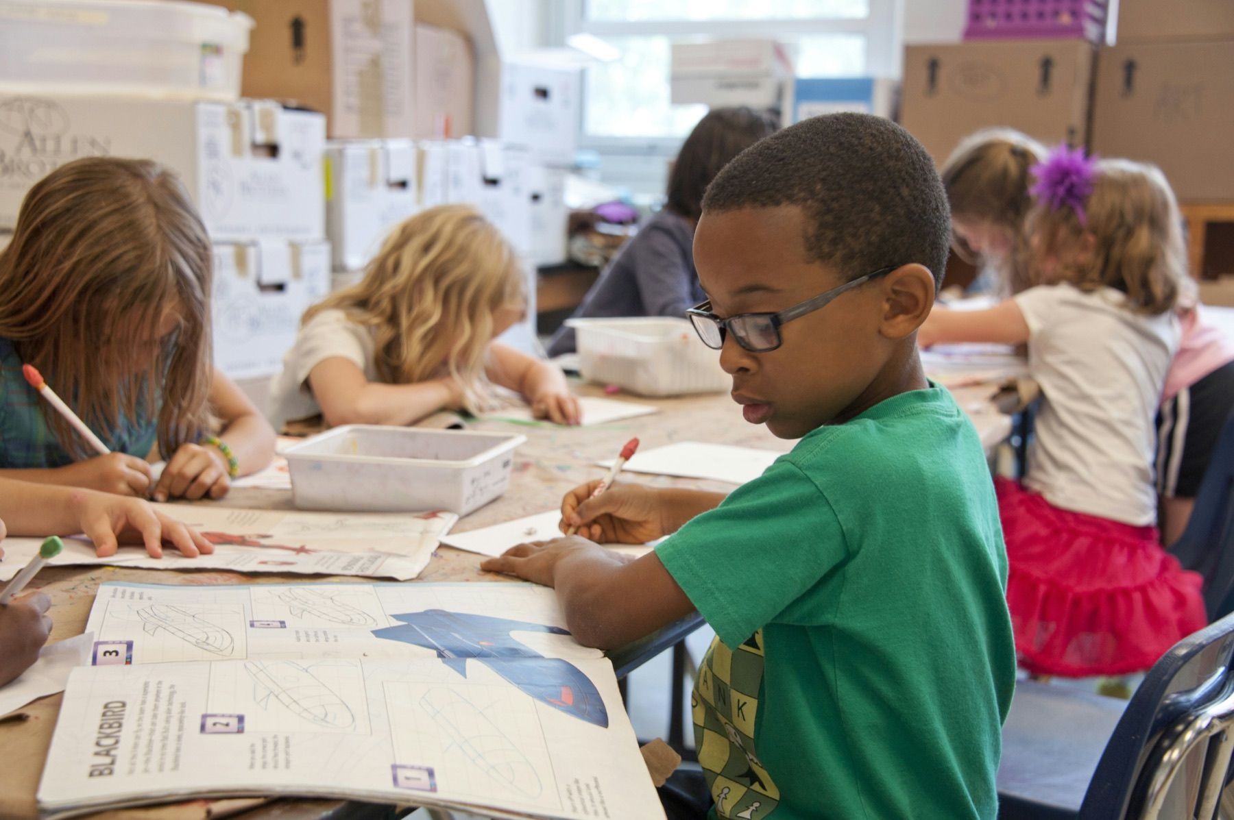 A schoolboy wearing glasses concentrates on a drawing while classmates work around the same table.